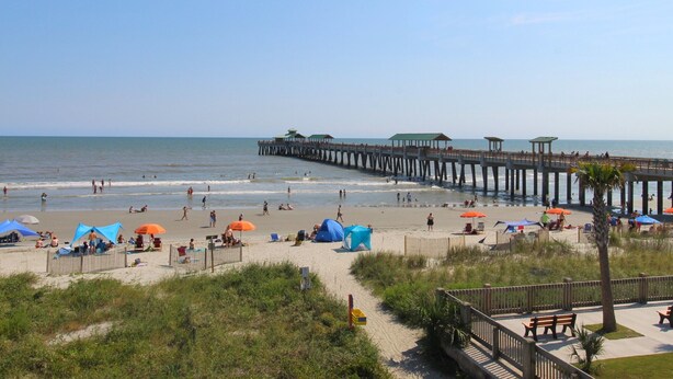 A beach with a wooden pier extending into the ocean, tents, and beachgoers. - Spacious Oceanfront and Pier Views~ Seaside Villas 202 (Folly Beach)