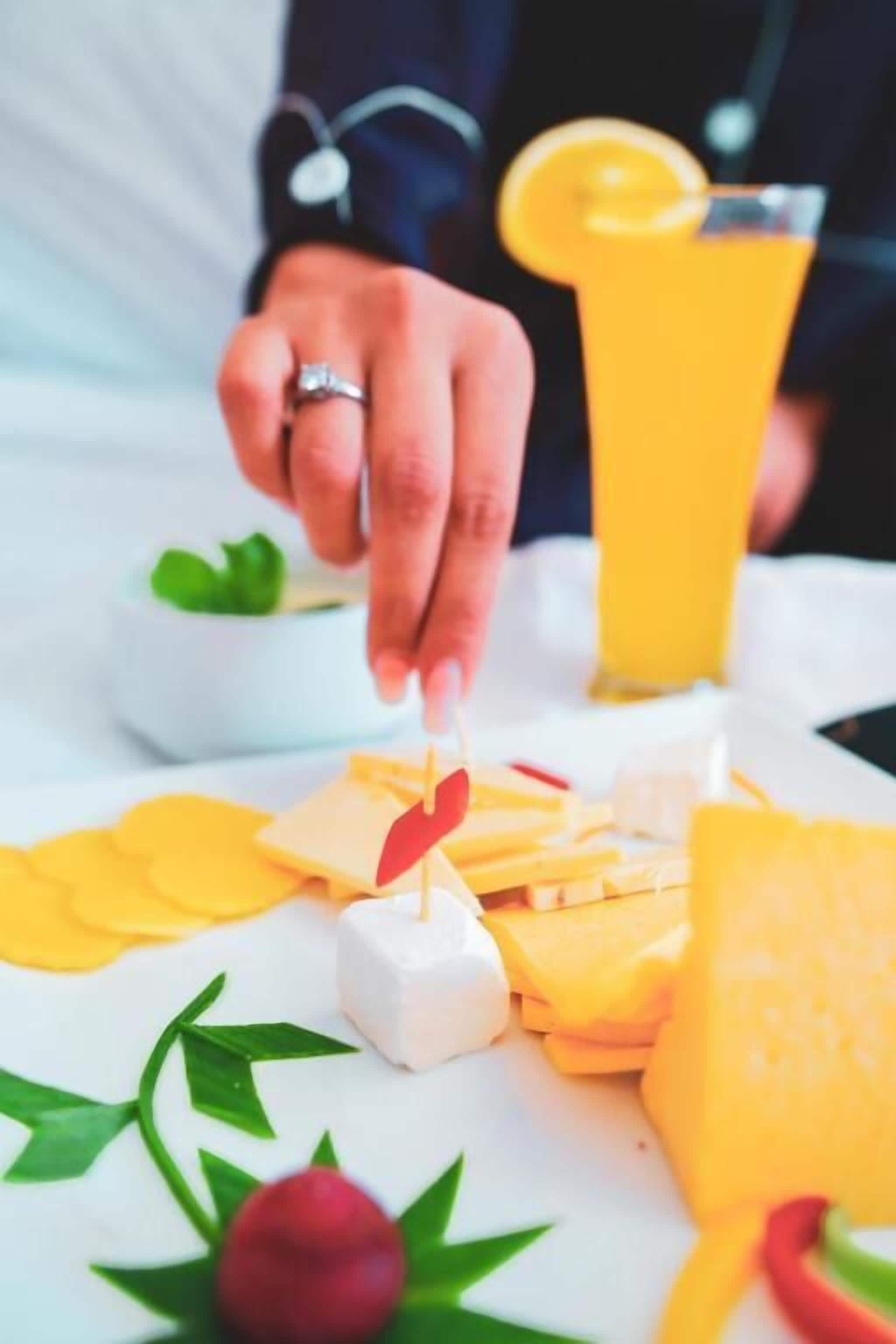 A person placing a red leaf-shaped decoration on a cheese platter with yellow cheese and a glass of orange juice.