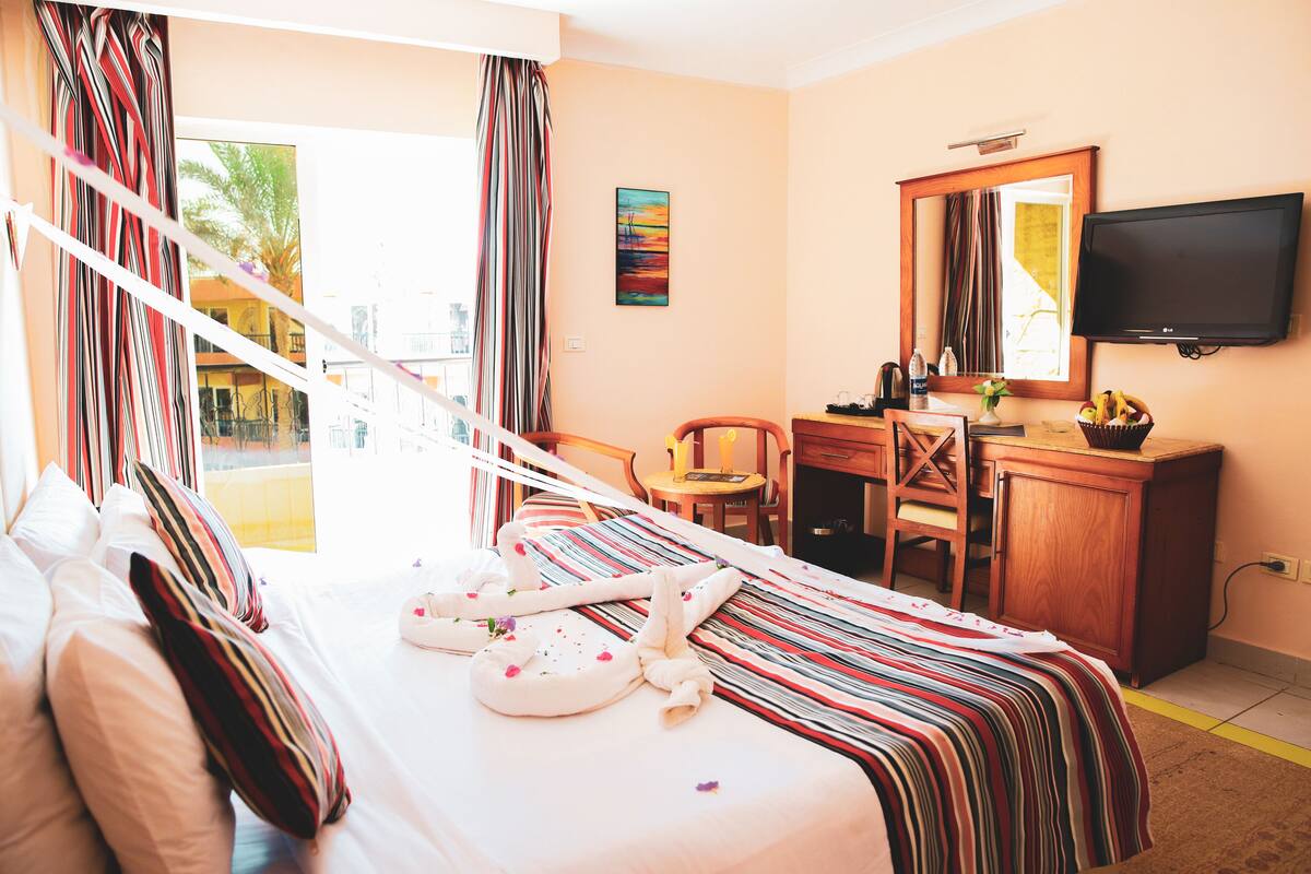 Habitación de hotel con cama, escritorio, silla, televisor y ventana con cortinas.