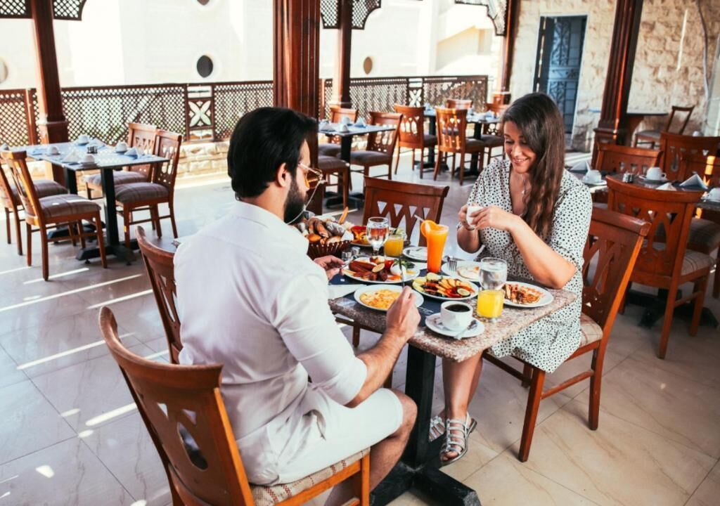 Una pareja cenando en un café al aire libre con varias mesas y sillas, piso de baldosas y vistas del entorno.