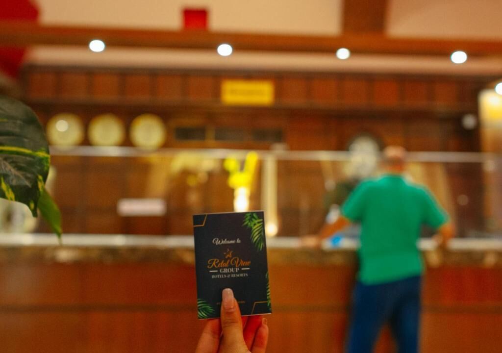 Una mano sosteniendo una tarjeta con el texto 'Royal Wine Group', un área de recepción con mostrador y una persona con camisa verde al fondo.