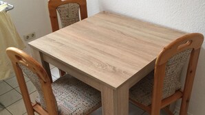 Ein Holzespeisesaal mit vier Stühlen, jeder mit einem gemusterten Kissen. Der Tisch steht auf einem Fliesenboden, im Hintergrund ist eine weiße Wand zu sehen.