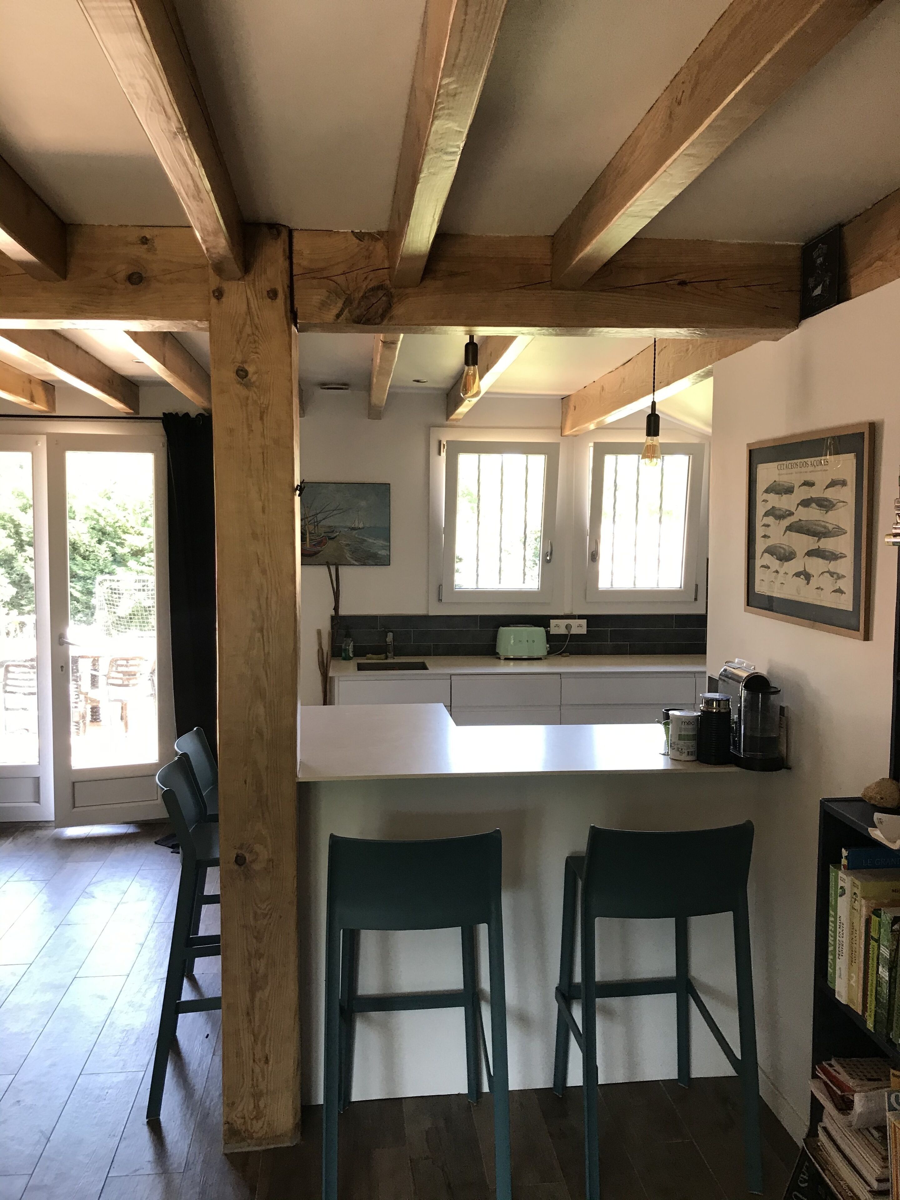 Eine Küche mit Holzbalken, einer weißen Arbeitsplatte, zwei Barhocker, einem Fenster mit Jalousien und einer gerahmten Illustration von Fischen an der Wand.