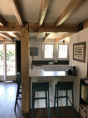 A kitchen with wooden beams, a white countertop, two bar stools, a window with blinds, and a framed fish illustration on the wall. - Maison Mango à Capbreton Bien Placée au Bouheben en Bordure de Foret (Capbreton)
