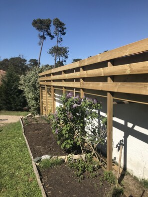 A wooden pergola structure with a garden bed beneath it, surrounded by a white wall and a grassy area. - Maison Mango à Capbreton Bien Placée au Bouheben en Bordure de Foret (Capbreton)
