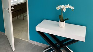 Una habitación con una pared verde azulado, una puerta blanca que da a un baño, una mesa blanca con un jarrón de flores blancas y una silla blanco y negro.