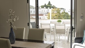 A balcony with white chairs and a table, a vase with flowers, and a view of buildings and trees. - Marina Luxor - Vilamoura (Vilamoura)