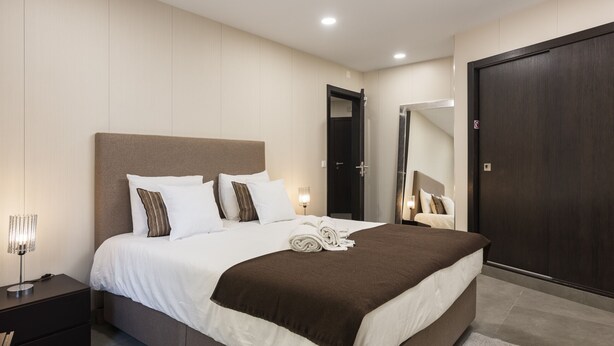 A modern hotel room with a large bed, a bedside table, a mirror, and a dark wooden door. - Marina Luxor - Vilamoura (Vilamoura)