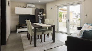A modern dining area with a table, chairs, and a vase of flowers. There is a sofa and a sliding glass door leading to an outdoor patio. - Marina Luxor - Vilamoura (Vilamoura)