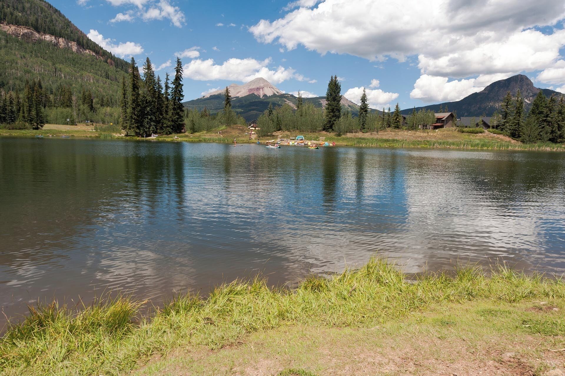 A calm lake with a mountain backdrop, surrounded by dense forest and a few buildings.