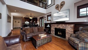 A spacious living room with a fireplace, a large flat-screen TV, and a high ceiling with a balcony.