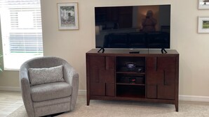 Ein Wohnzimmer mit Flachbildfernseher auf einem Holzboden, einem grauen Sessel und einem gerahmten Bild an der Wand.