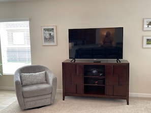 A living room with a flat-screen TV on a wooden stand, a grey armchair, and a framed picture on the wall. - The Sweet Spot Between Comfort & Adventure (The Villages)