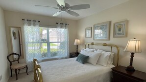 Ein Schlafzimmer mit Bett, Deckenventilator, Fenster mit Vorhängen, Stuhl, Nachttisch mit Lampe und gerahmten Bildern an der Wand.