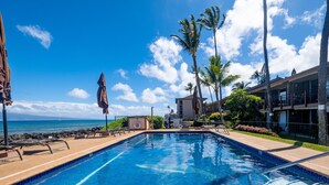 A swimming pool with lounge chairs and umbrellas, a view of the ocean, and a building with balconies.