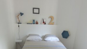 Ein ordentlich bezogenes Bett mit gestreifter Bettdecke und zwei Kissen, ein kleiner Nachttisch mit einer Lampe sowie ein Wandregal mit verschiedenen Gegenständen.