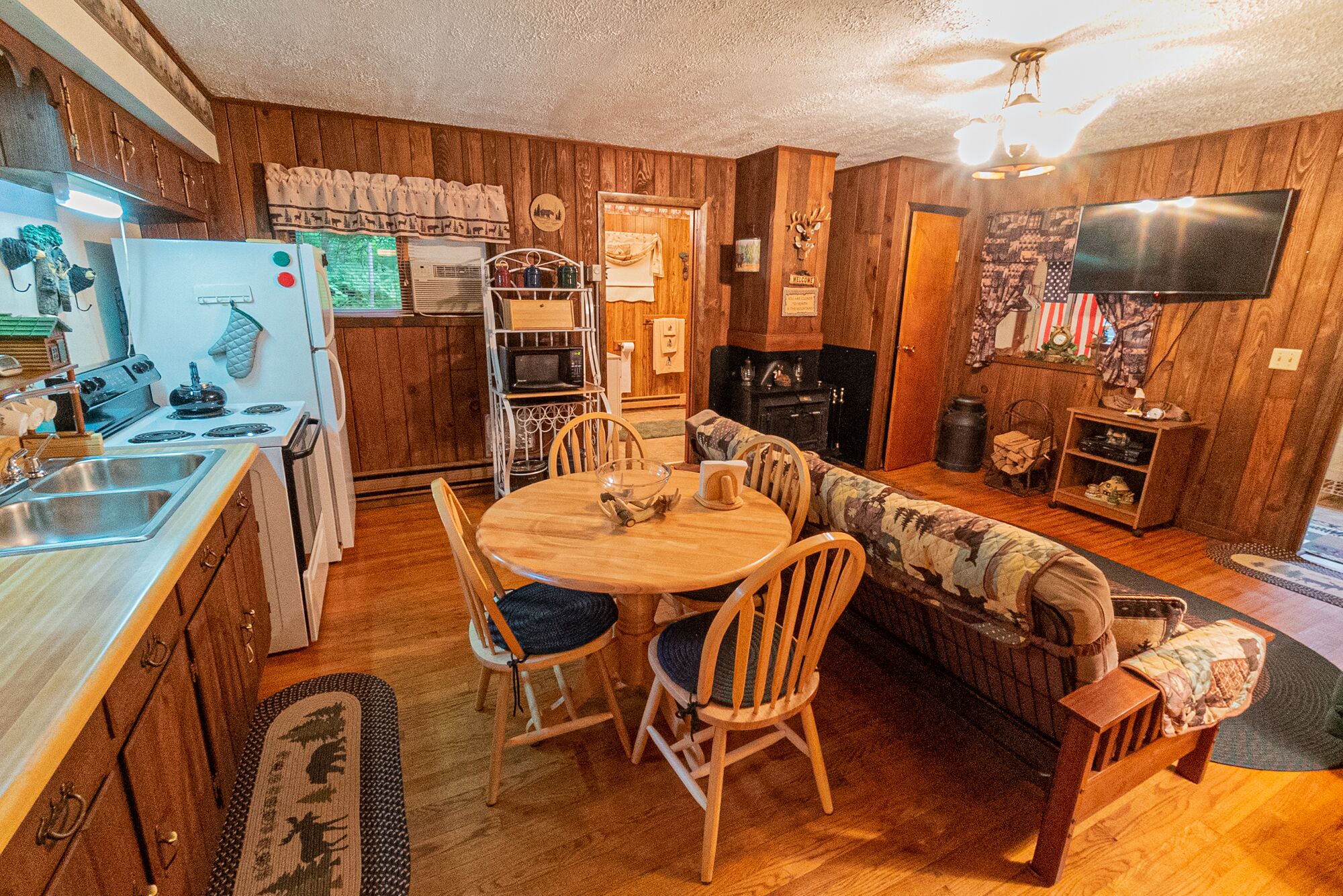 A cozy cabin-style room with a wooden interior, a dining table with chairs, a sofa, a kitchen area with a stove and sink, and a television mounted on the wall.