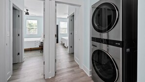 Pesuhuoneessa on kaksi päällekkäin asennettua, edestä täytettävää pesu- ja kuivauskonetta, kattotuuletin sekä käytävä, joka johtaa makuuhuoneeseen, jossa on sänky ja ikkuna, jossa on sälekaihtimet.