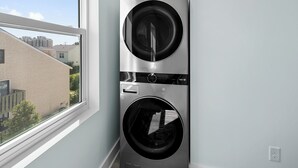 A modern laundry room with two stacked front-loading washing machines, a window with a view of buildings, and a white wall. - MAIN EVENT HOME 20beds/19baths CINEMA+AMENITIES+Pool ONLY 350ft to beach!!! (Myrtle Beach)