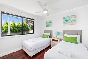 A bedroom with two beds, a ceiling fan, a window with a view of trees, and two wall-mounted paintings. - The Tomaree Upper Apartment (Shoal Bay)