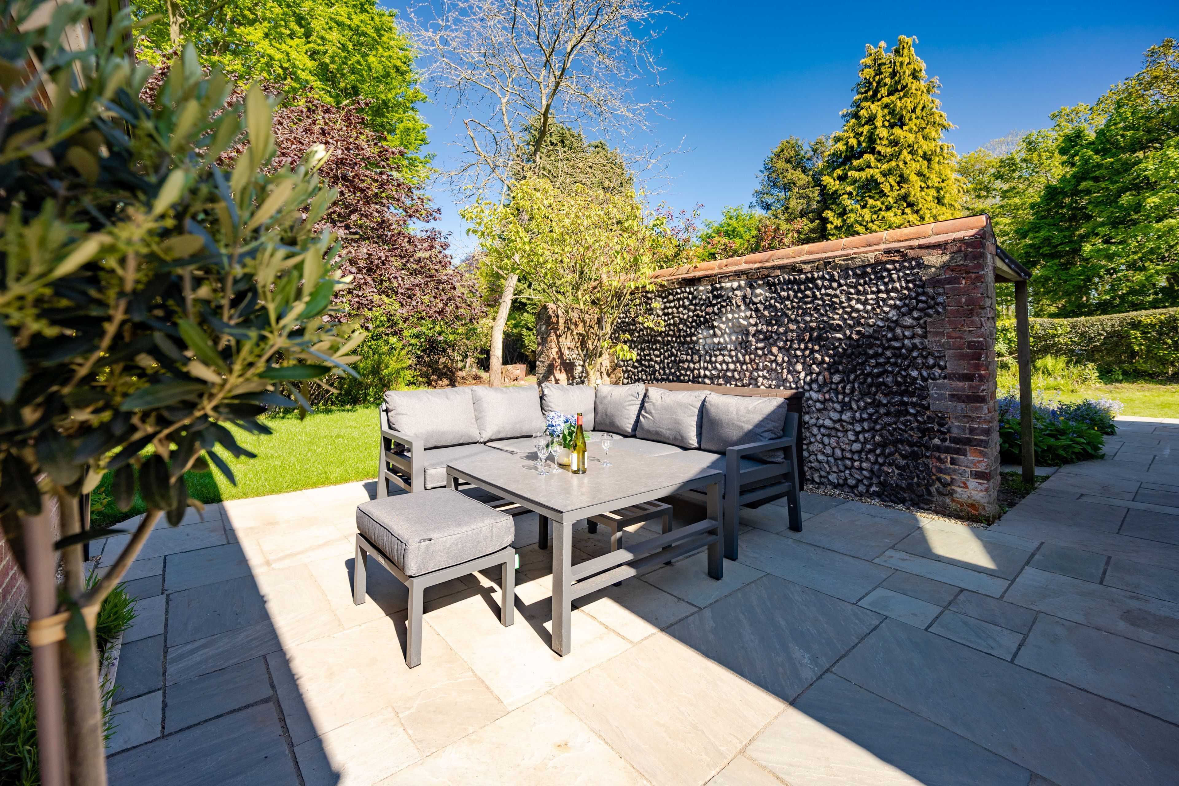 An outdoor patio with a stone wall, a grey sofa set, and a table with a bottle and glasses.