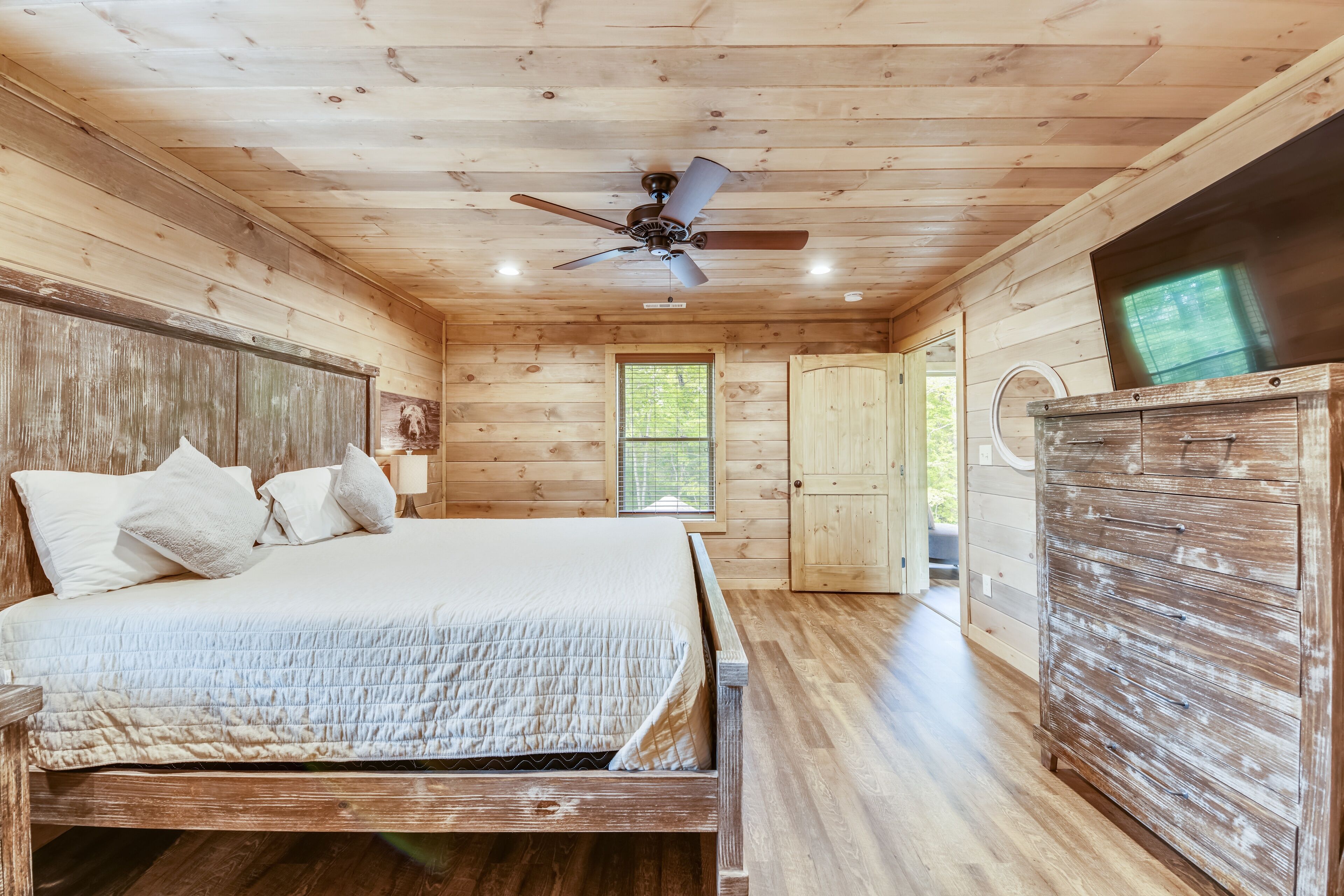Un dormitorio de cabaña de madera con cama, cómoda, ventilador de techo y ventana.