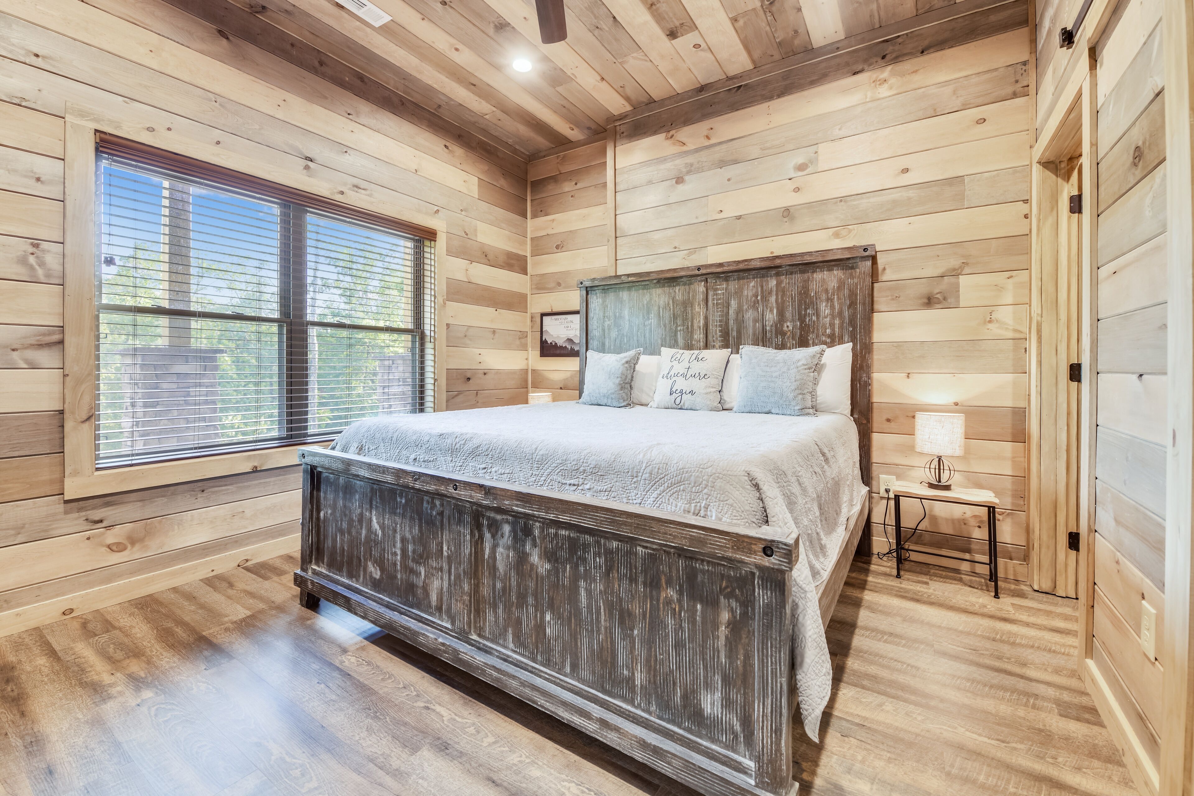 Un dormitorio con paneles de madera, una cama grande, una mesita de noche pequeña y un ventanal con persianas.