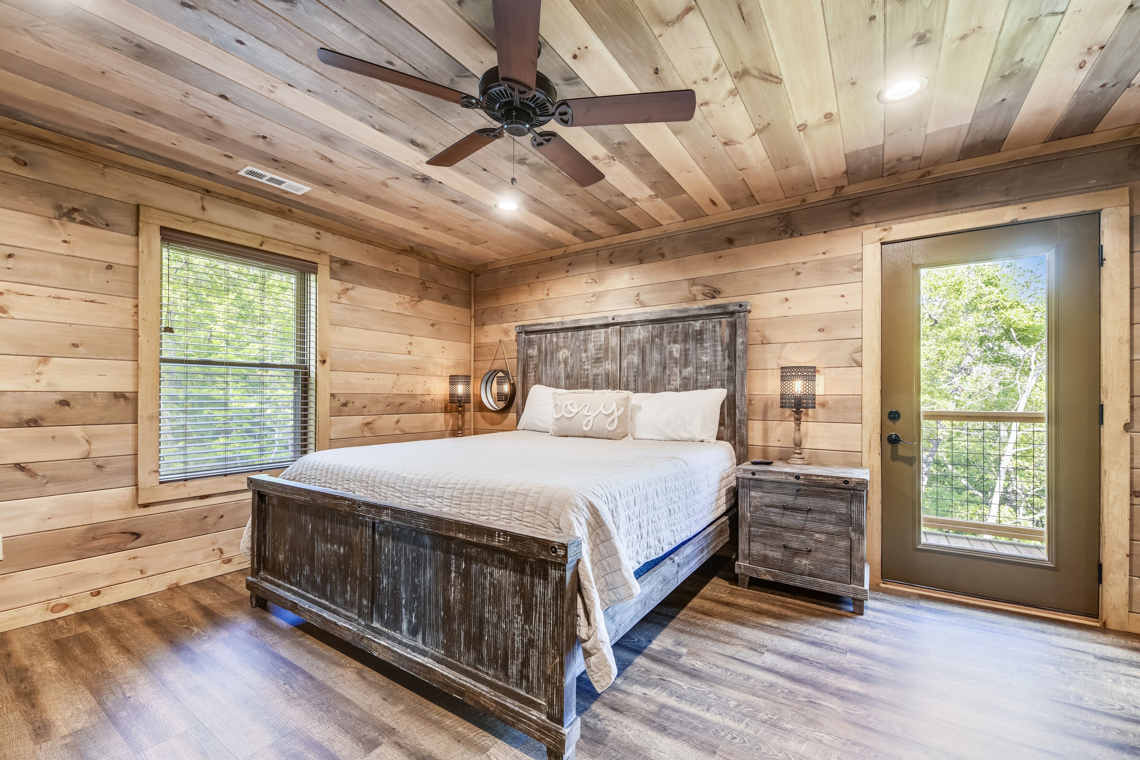 A wooden cabin bedroom with a bed, a nightstand, a ceiling fan, and a door leading outside.