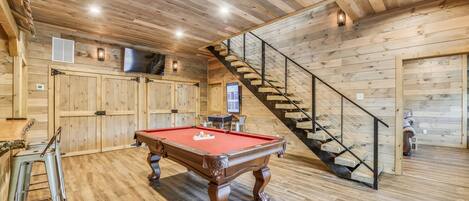 A wooden interior with a pool table, chairs, a staircase, and a TV.