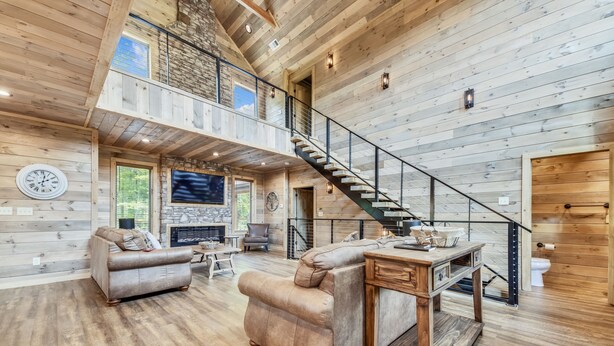 A spacious living area with a wooden floor, a leather sofa, a coffee table, and a fireplace. A staircase with a glass railing leads to an upper level.