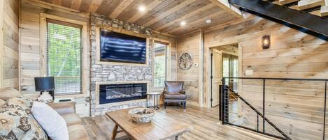 A cozy log cabin interior with a stone fireplace, a comfortable sofa, a wooden coffee table, and a staircase.