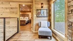 A wooden cabin interior with a cozy bed, a small seating area, and a large window offering a view of trees.
