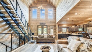 A spacious interior with a high ceiling, wooden walls, a staircase, a living area with a sofa and a coffee table, and a kitchen area.