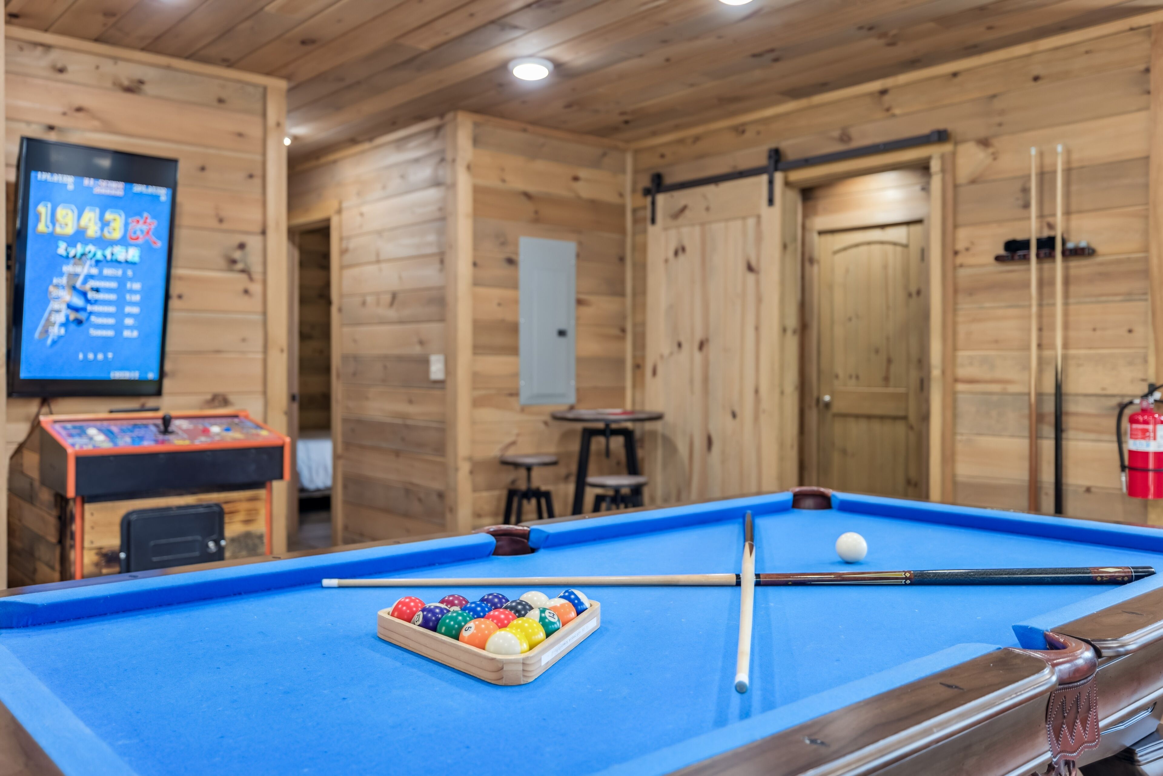 A pool table with balls and cues, a television displaying information, and wooden walls.