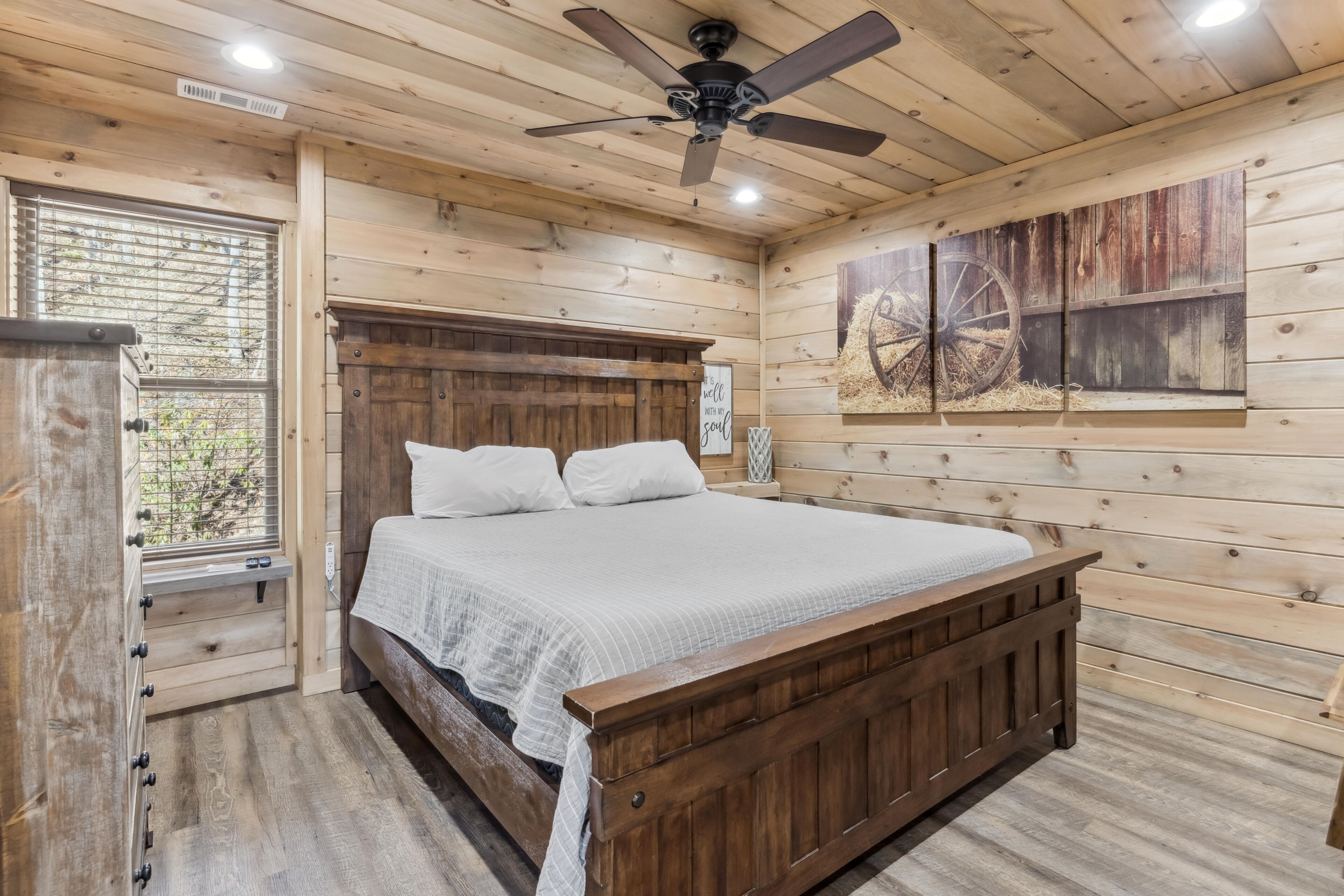 A wooden bedroom with a bed, a ceiling fan, a window, and a wall-mounted artwork.