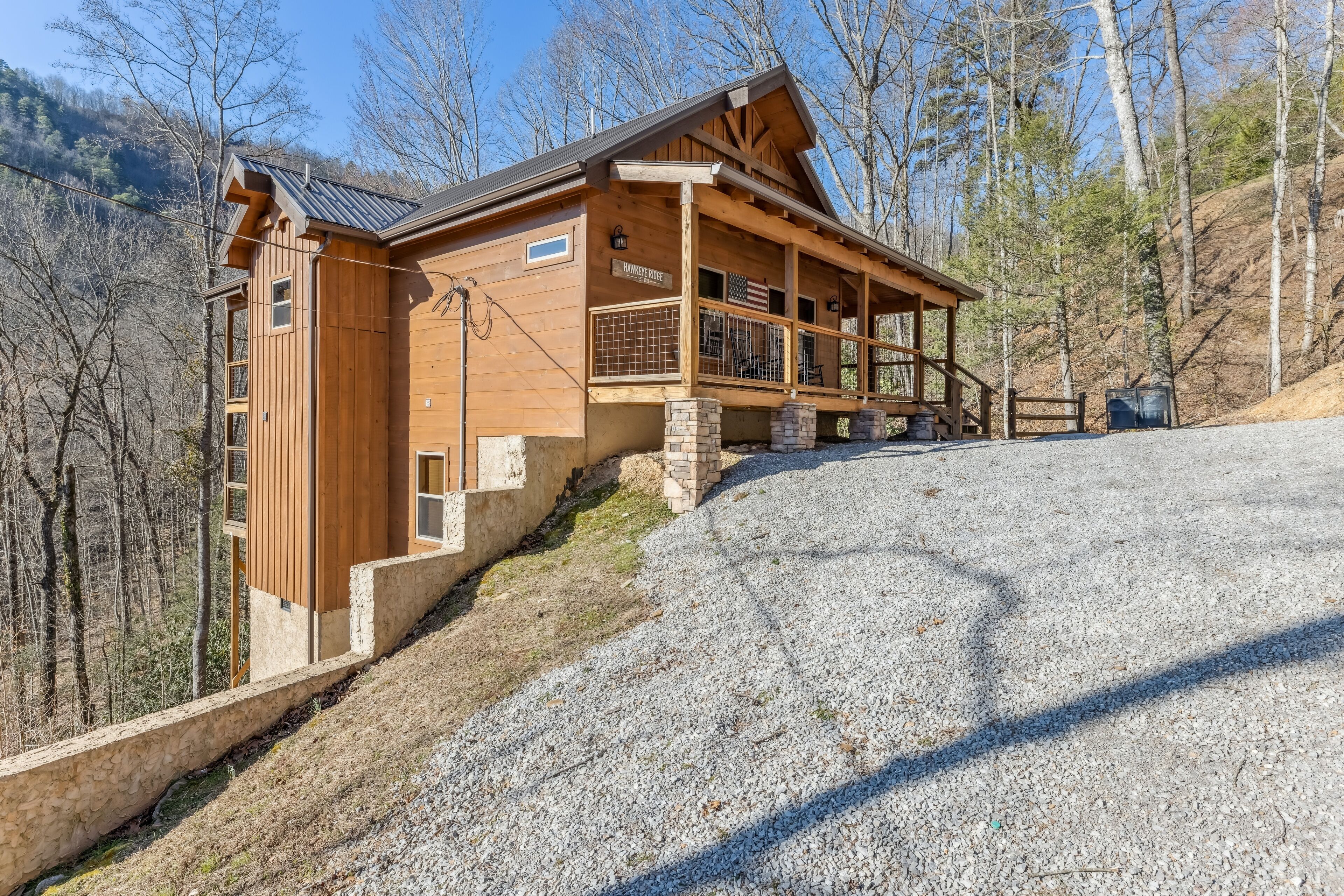A wooden cabin with a porch, stone foundation, and a view of the surrounding forest.