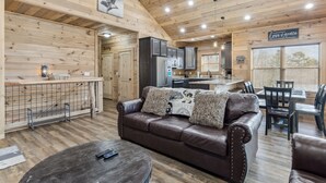 Un interior de cabaña acogedor, con techo de madera, un sofá de cuero marrón con cojines estampados, una mesa de comedor con sillas y una zona de cocina con armarios oscuros y un refrigerador.