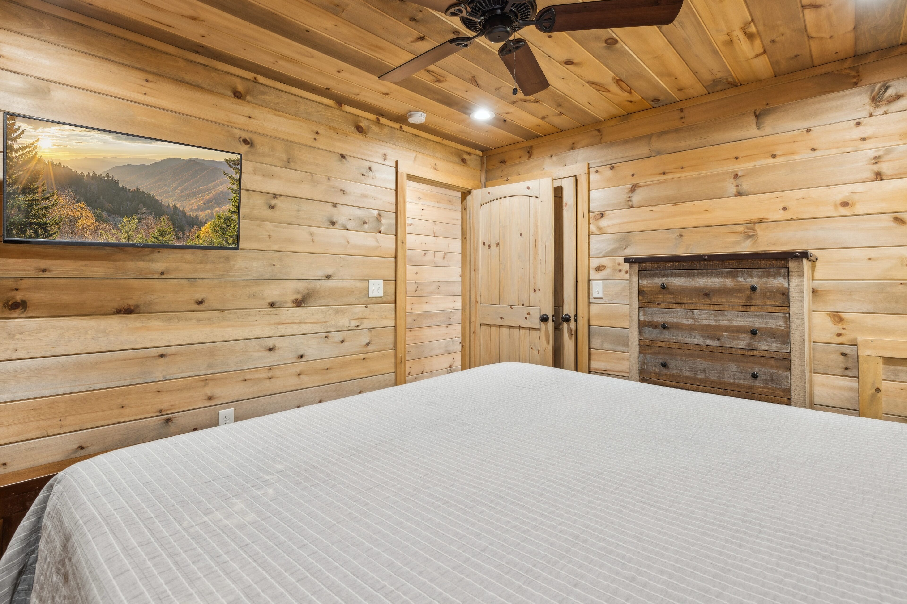 A wooden cabin room with a bed, a ceiling fan, a fireplace, and a painting of a mountain landscape.