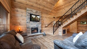 A cozy cabin interior with a stone fireplace, leather sofas, a TV mounted on the wall, and a wooden staircase.