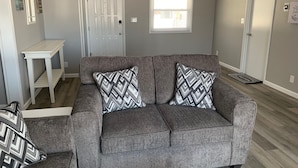A grey sofa with patterned cushions, a white side table, and a window with blinds. - Lake Front home near Sister Lakes area.  Pontoon rental available. (Decatur)