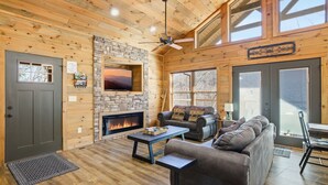 A cozy cabin interior with a stone fireplace, wooden walls, a ceiling fan, and a large window. - Brand New Freedom Cabin with Large Deck by RedAwning (Sevierville)