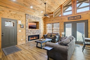 A cozy cabin interior with a stone fireplace, wooden walls, a ceiling fan, and a large window. - Brand New Freedom Cabin with Large Deck by RedAwning (Sevierville)