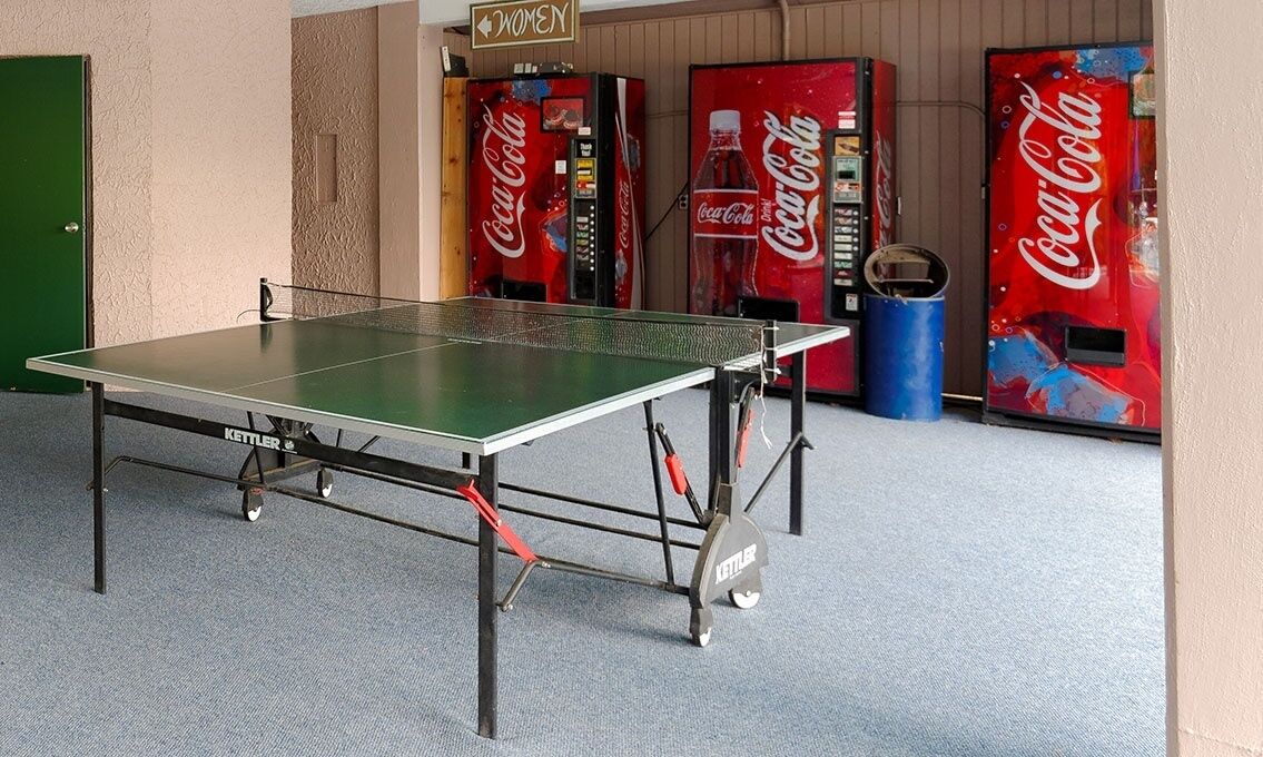 A ping pong table in a room with a green door and walls, and three Coca-Cola vending machines.