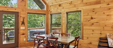 A wooden cabin with a dining area featuring a table and chairs, large windows, and a view of greenery outside.