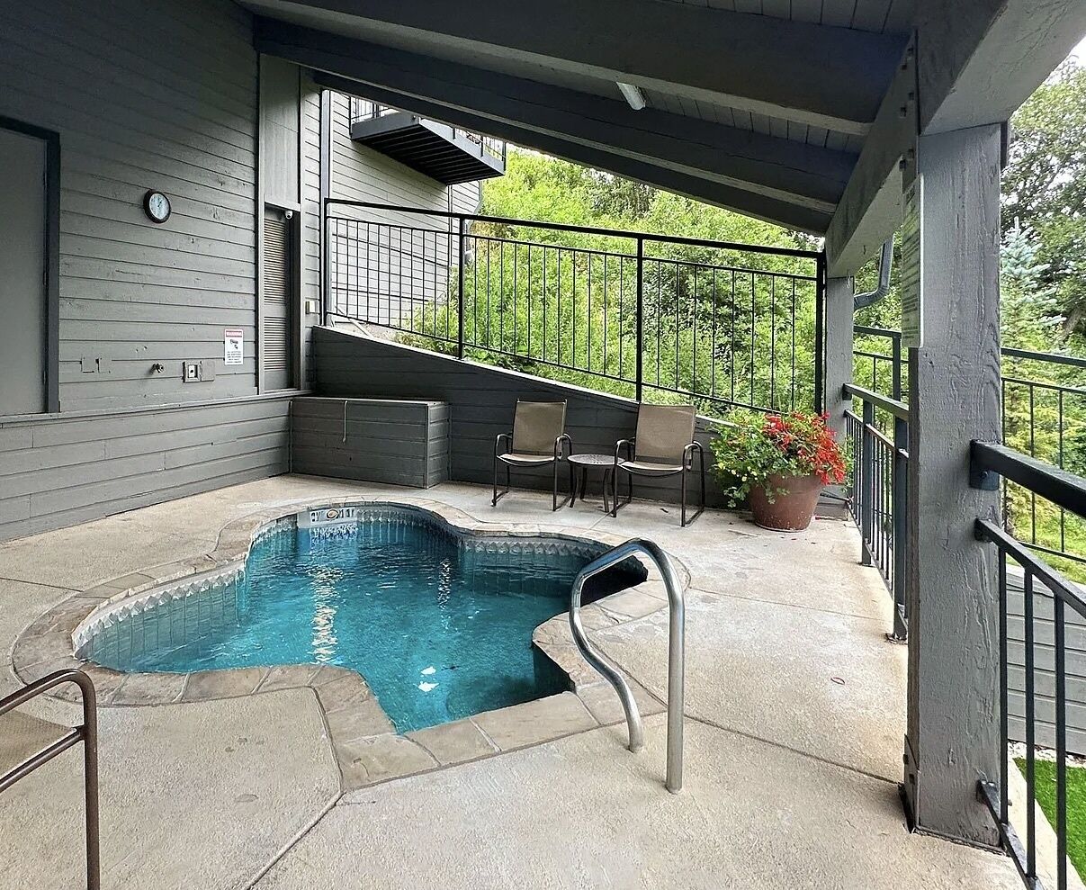 A covered outdoor swimming pool with a metal ladder, adjacent seating area, and a staircase leading to an upper level.