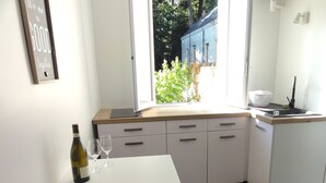 A kitchen with a white table, a wine bottle, and two glasses. There is a window with a view of trees and a building outside. - L'etoile Loire - Un Jardin en Ville ! (Saumur)