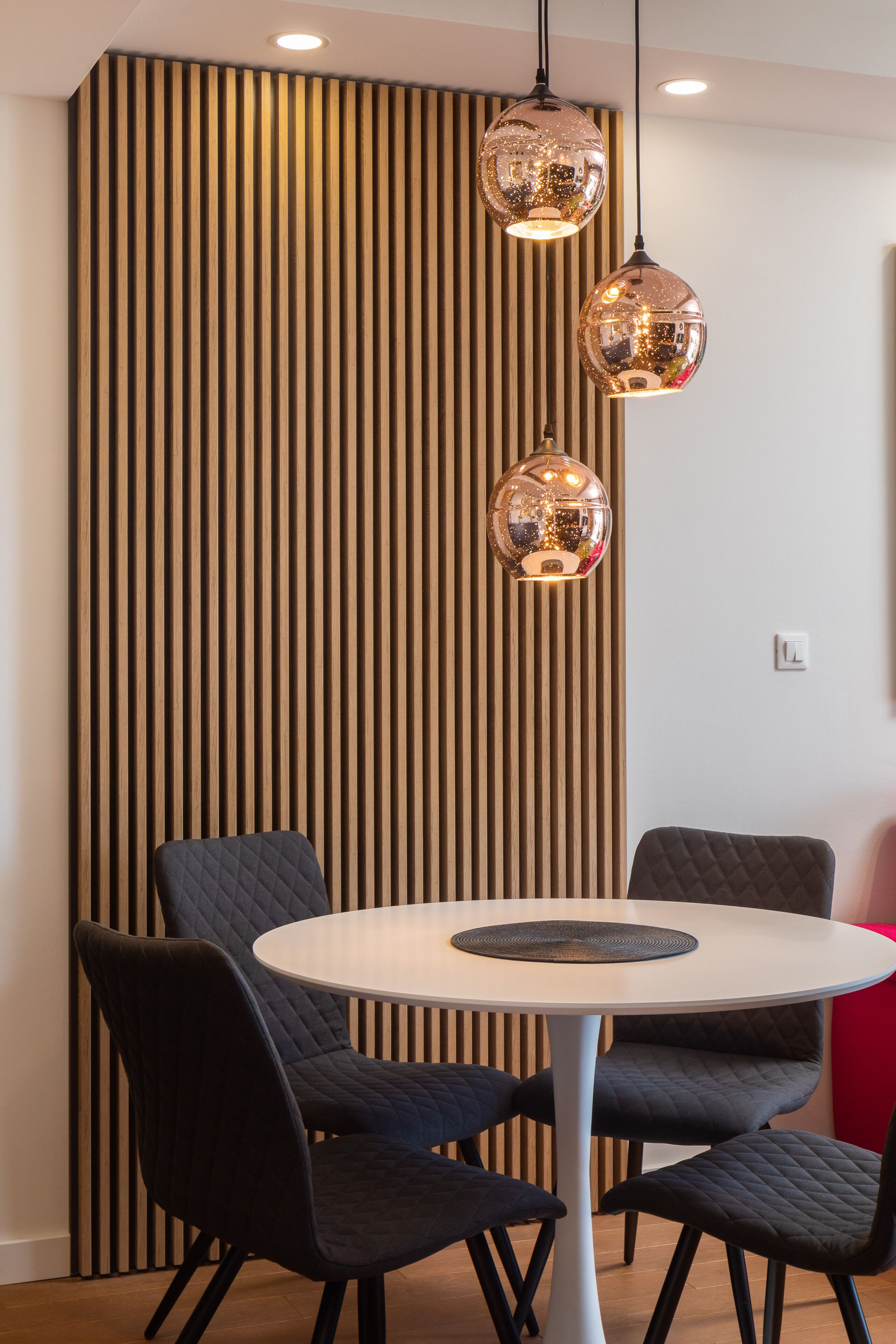 Un comedor moderno con una mesa redonda y cuatro sillas, una pared con listones de madera y dos lámparas colgantes esféricas.