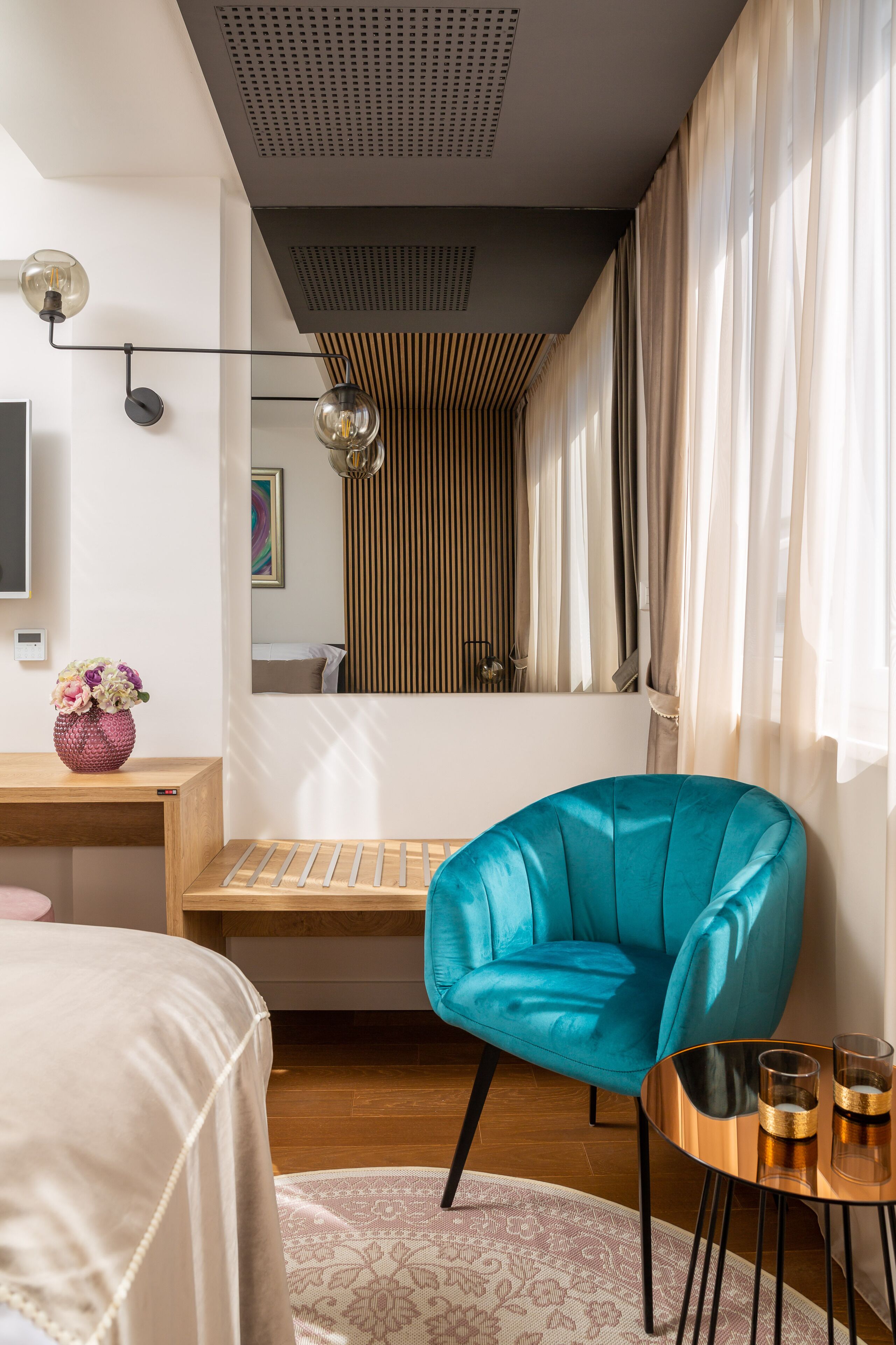 Ett modernt hotellrum med ett skrivbord i trä, en turkosfärgad fåtölj, ett litet runt bord med glas och ett stort fönster med gardiner.