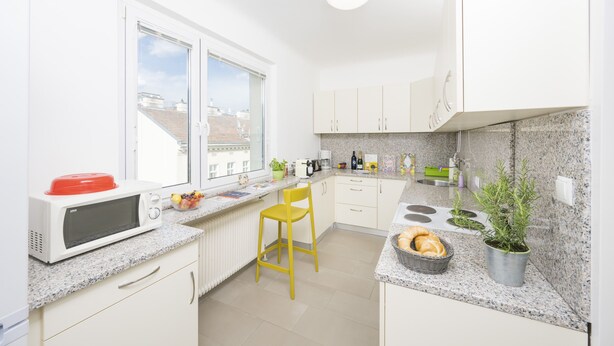 Una cocina moderna con armarios blancos, un microondas rojo, una silla amarilla, una ventana con vistas a edificios y una planta en maceta.