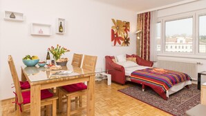 Un apartamento moderno con comedor, una cama con manta colorida, un sofá rojo y un amplio ventanal con vistas a edificios.